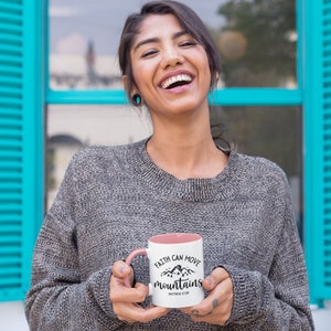May include: A white ceramic mug with a pink interior and handle, featuring a black graphic of mountains and pine trees with the text "FAITH CAN MOVE mountains" and "MATTHEW 17:20" printed in a decorative black script and sans-serif font.