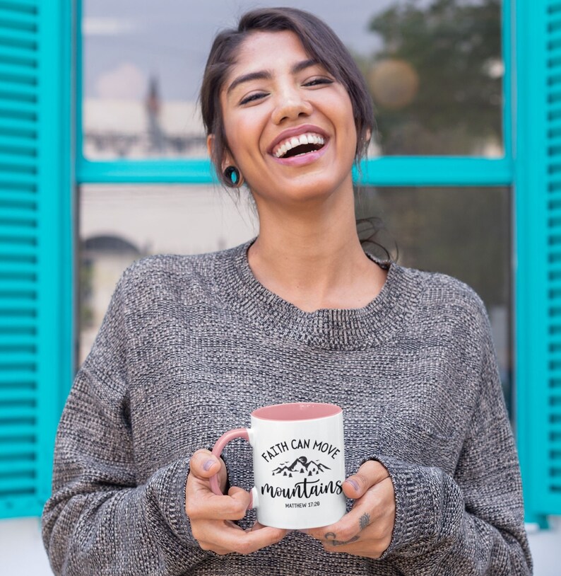 May include: A white ceramic mug with a pink interior and handle, featuring a black graphic of mountains and pine trees with the text "FAITH CAN MOVE mountains" and "MATTHEW 17:20" printed in a decorative black script and sans-serif font.