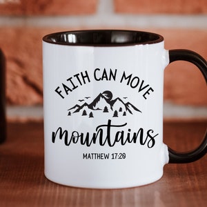 May include: A white ceramic mug with a black handle and interior rim. The front features a black graphic of mountains, trees, and birds with the text "FAITH CAN MOVE mountains" and "MATTHEW 17:20" printed in a combination of serif and script fonts.