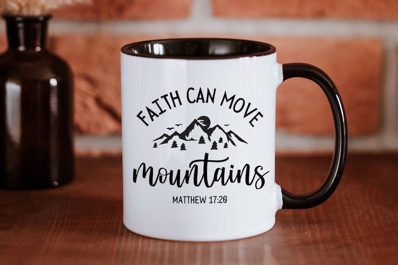 May include: A white ceramic mug with a black handle and interior rim. The front features a black graphic of mountains, trees, and birds with the text "FAITH CAN MOVE mountains" and "MATTHEW 17:20" printed in a combination of serif and script fonts.