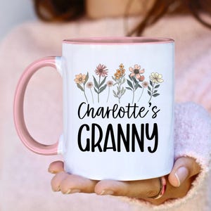 Op de afbeelding: Witte keramische mok met een roze handvat en binnenkant. De mok heeft een bloemenontwerp met verschillende bloemen en de tekst "Charlotte's GRANNY" in het zwart. De mok wordt door een persoon vastgehouden.