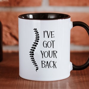 Op de afbeelding: Een witte keramische mok met een zwarte binnenkant en een zwart handvat. De voorzijde van de mok toont een zwarte afbeelding van een menselijke ruggengraat links van de zwarte tekst, I'VE GOT YOUR BACK, in een eenvoudig schreeflettertype.
