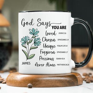 May include: White ceramic mug with a black handle and rim, featuring the text "God Says You Are" and inspirational words. The mug has a floral design with blue flowers and green leaves. It sits on a wooden coaster.