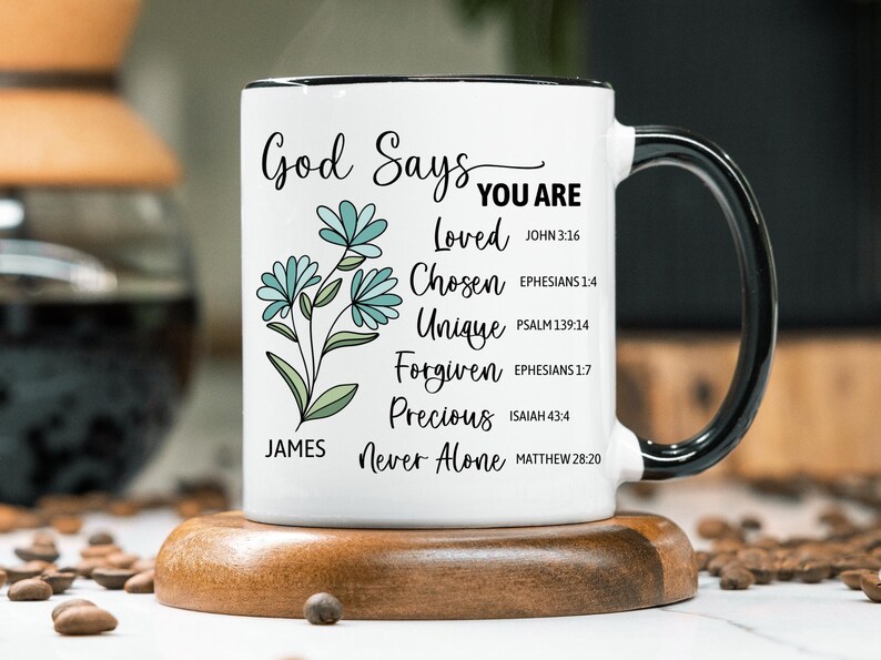 May include: White ceramic mug with a black handle and rim, featuring the text "God Says You Are" and inspirational words. The mug has a floral design with blue flowers and green leaves. It sits on a wooden coaster.