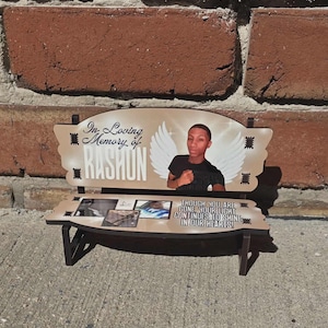 Könnte beinhalten: Ein dekoratives bankförmiges Denkmal mit einem Foto einer Person, Engelsflügeln und dem Text "In Loving Memory of RASHON". Die Bank enthält auch den Text "THOUGH YOU ARE GONE YOUR LIGHT CONTINUES TO SHINE IN OUR HEARTS!"