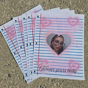Puede incluir: Una pila de hojas de papel rayado con un recorte en forma de corazón que muestra a una persona con gafas. Las hojas tienen líneas azules, márgenes rojos y diseños de corazón rosa. El texto "Forever yours baby" está impreso en la parte inferior.