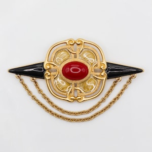 May include: A gold-toned brooch with a central red oval stone. The brooch features a decorative design with black accents and two gold chains hanging below. The brooch is a vintage accessory.