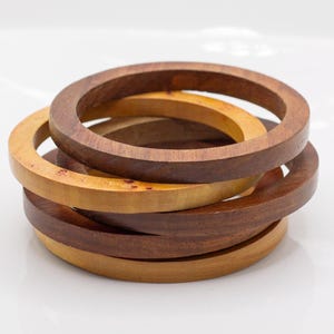May include: A stack of four circular wooden bangles featuring alternating light and dark brown wood tones. The bracelets have a smooth, polished finish and a simple, rounded band design, showcasing natural wood grain patterns throughout the set.