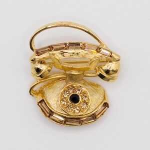 May include: A gold-tone metal brooch shaped like a vintage rotary telephone. The piece features a textured finish, a central black stone surrounded by small clear crystals on the dial, and baguette-cut crystals along the base and handset.