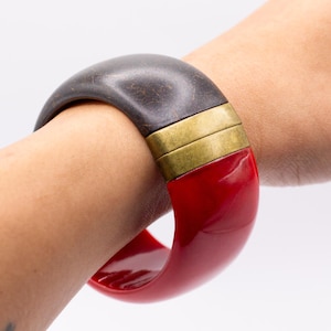 May include: A wide cuff bracelet featuring a dark brown textured segment and a glossy red segment, connected by two horizontal brushed brass-toned metal bands. The bracelet has a smooth, curved profile and a substantial, solid construction.