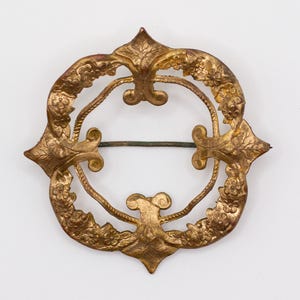 May include: A circular gold-toned metal brooch featuring an ornate, embossed floral pattern along the outer rim. The design includes four symmetrical, leaf-like protrusions extending from the frame, with a thin, horizontal metal pin across the center.