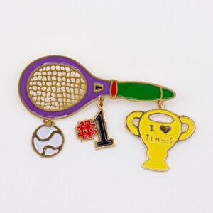 May include: A gold-tone metal brooch featuring a purple tennis racket with a green handle, a white tennis ball, a red and black number one charm, and a yellow trophy charm with the text I heart TENNIS engraved on the front surface.