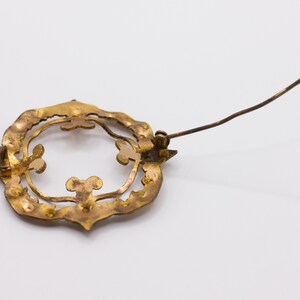 May include: A gold-toned metal brooch featuring a circular, openwork frame with decorative scalloped edges and interior clover-like accents. The back of the piece displays a long, thin metal pin mechanism attached to the frame for fastening.