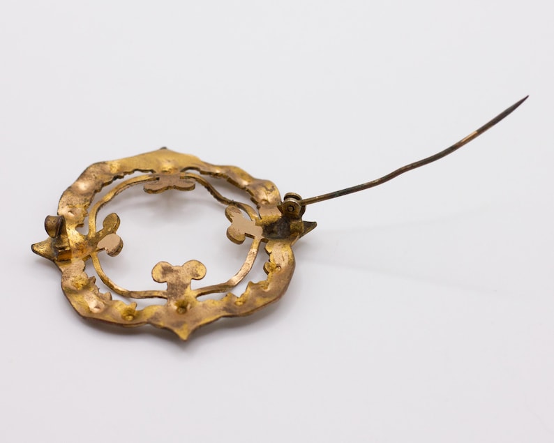 May include: A gold-toned metal brooch featuring a circular, openwork frame with decorative scalloped edges and interior clover-like accents. The back of the piece displays a long, thin metal pin mechanism attached to the frame for fastening.