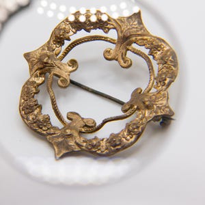 May include: A circular gold-toned metal brooch featuring an intricate openwork design with embossed floral and leaf motifs. The piece includes a thin horizontal pin mechanism attached to the back, visible through the center of the decorative frame.