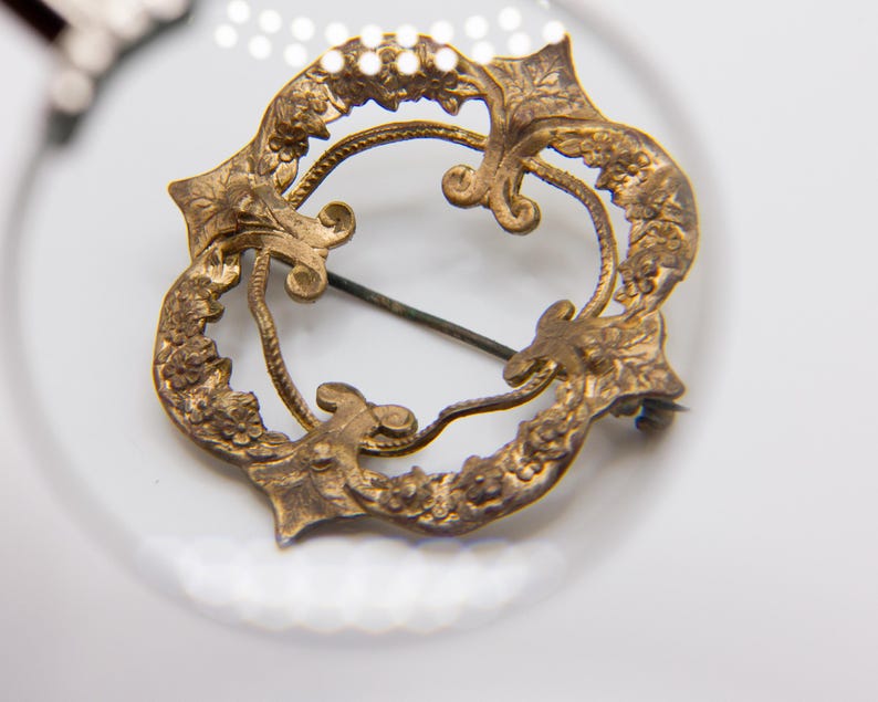 May include: A circular gold-toned metal brooch featuring an intricate openwork design with embossed floral and leaf motifs. The piece includes a thin horizontal pin mechanism attached to the back, visible through the center of the decorative frame.