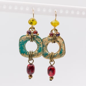 May include: Dangle earrings featuring a square, textured teal and bronze metal frame. Each earring is accented with a yellow faceted bead at the top, small multicolored beads in the center, and a single smooth, glossy red bead at the bottom.