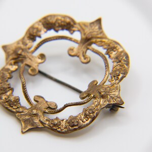 May include: A circular brass brooch featuring an intricate embossed floral and leaf pattern along the outer rim. The design includes four pointed accents and a central open space with a thin metal pin extending across the diameter of the piece.