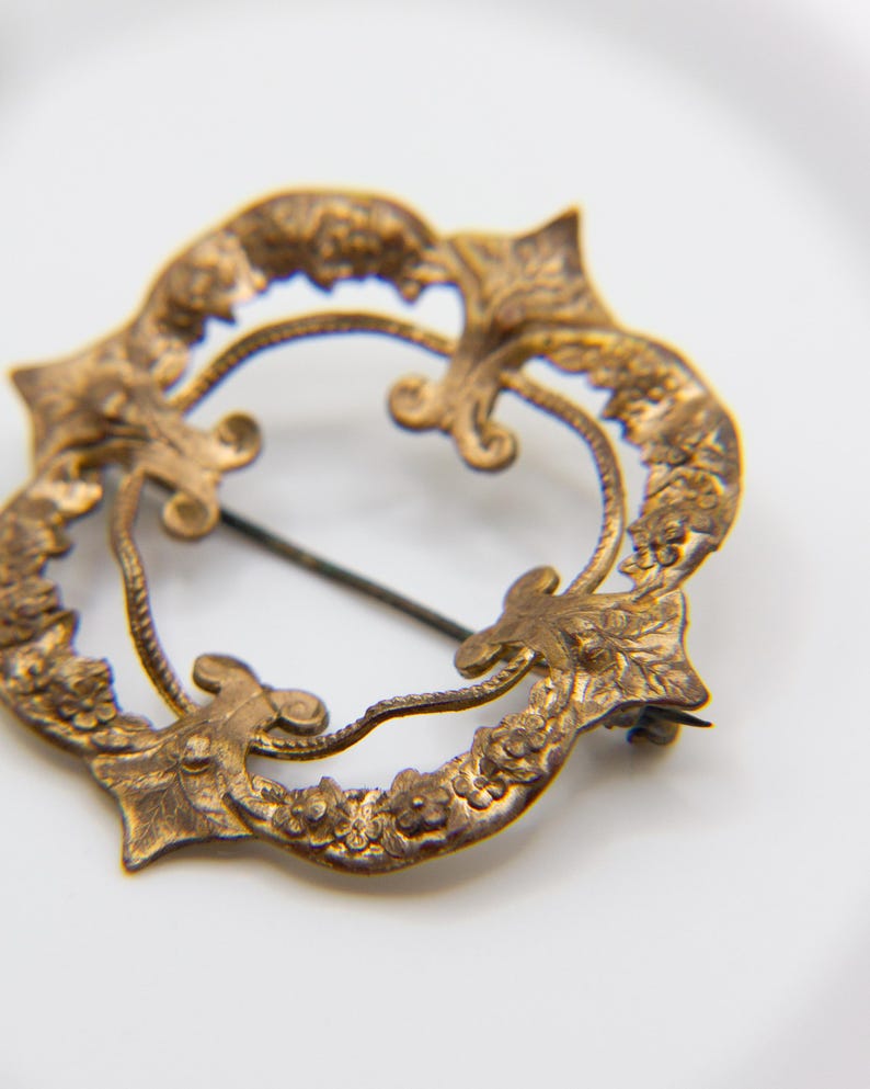May include: A circular brass brooch featuring an intricate embossed floral and leaf pattern along the outer rim. The design includes four pointed accents and a central open space with a thin metal pin extending across the diameter of the piece.