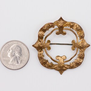 May include: A circular gold-toned metal brooch featuring an intricate embossed floral pattern and a central open space with a horizontal pin mechanism, positioned next to a United States quarter coin for scale to show its relative size.