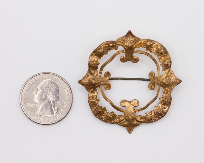 May include: A circular gold-toned metal brooch featuring an intricate embossed floral pattern and a central open space with a horizontal pin mechanism, positioned next to a United States quarter coin for scale to show its relative size.