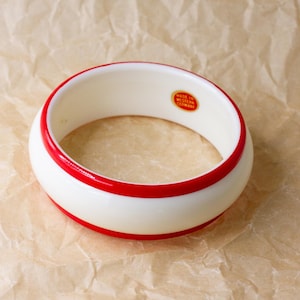 May include: A thick, circular bangle bracelet featuring a smooth, off-white base with two vibrant red bands encircling the top and bottom edges. Inside the bracelet, a small, oval-shaped sticker reads Made in Western Germany in red text.