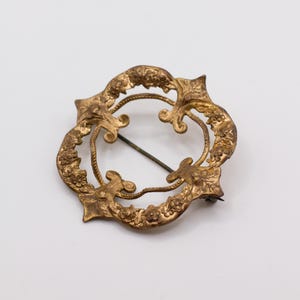 May include: A circular gold-toned metal brooch featuring an intricate embossed floral pattern along the outer rim. The design includes four pointed fleur-de-lis style accents and a thin, dark metal pin mechanism visible across the open center.