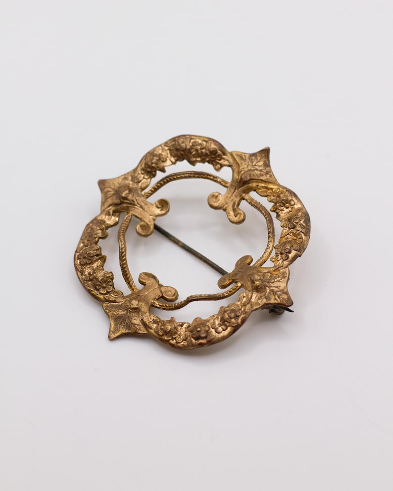 May include: A circular gold-toned metal brooch featuring an intricate embossed floral pattern along the outer rim. The design includes four pointed fleur-de-lis style accents and a thin, dark metal pin mechanism visible across the open center.