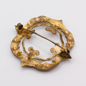 May include: A circular gold-toned metal brooch featuring an openwork design with scalloped edges and interior clover-like motifs. The back of the piece displays a thin metal pin mechanism with a hinge and a simple hook closure for attachment.