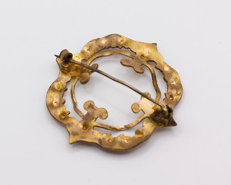 May include: A circular gold-toned metal brooch featuring an openwork design with scalloped edges and interior clover-like motifs. The back of the piece displays a thin metal pin mechanism with a hinge and a simple hook closure for attachment.