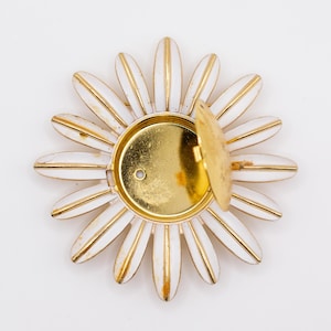 May include: A circular brooch shaped like a daisy with white enamel petals and gold-toned metal edges. The center features a small, round, hinged gold-toned locket compartment that is partially open, revealing a smooth, metallic interior surface.