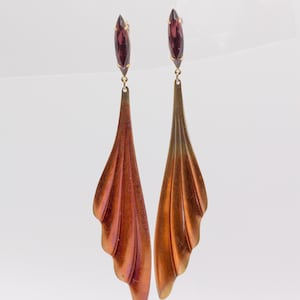 May include: A pair of long drop earrings featuring a marquise-cut deep purple gemstone at the top, connected to a large, fan-shaped metal pendant with a gradient copper and bronze finish and a series of vertical, curved ridges along the surface.
