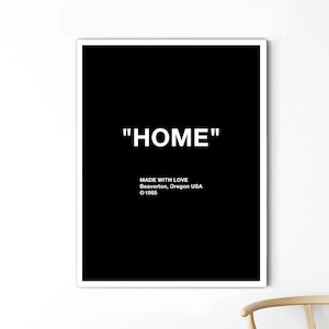 May include: A framed black and white art print with the word "HOME" in bold white letters. Below the word are the words "MADE WITH LOVE" and the location "Beaverton, Oregon USA" and the year "© 1985". The print is displayed on a white wall.