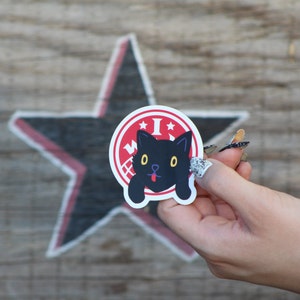 May include: A red and white sticker with a black cat illustration. The cat has yellow eyes and a red tongue sticking out. The sticker says "I Woof" in a white circle.