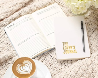 Könnte beinhalten: Ein weißes Notizbuch mit dem goldenen Schriftzug "THE LOVER'S JOURNAL", ein schwarzer Stift und ein offenes Notizbuch mit Text. Eine Kaffeetasse mit Latte Art und ein Strauß weißer Blumen sind ebenfalls vorhanden.