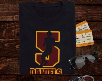 May include: Black t-shirt with a maroon and gold football player graphic. The graphic is a silhouette of a player inside a large letter "S". The name "DANIELS" is printed below the graphic. Sunglasses, tickets, and a car key are also in the image.