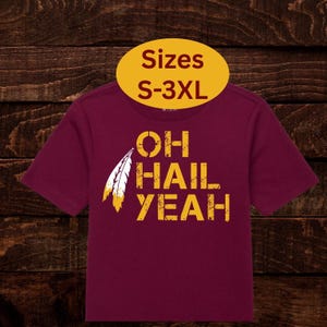 May include: Burgundy t-shirt with the text "OH HAIL YEAH" in yellow, featuring a feather graphic. A yellow oval above the shirt reads "Sizes S-3XL". The shirt has short sleeves.