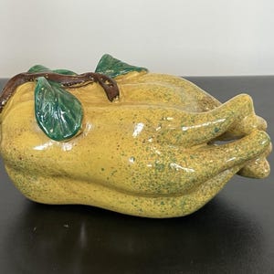 May include: A glazed ceramic sculpture shaped like a whole roasted chicken with a glossy yellow finish, speckled with green dots. The piece features a brown stem-like detail on top and green leaves, with the legs tucked toward the center.