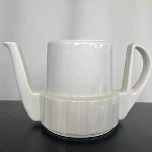 May include: A white ceramic teapot with a cylindrical body and a spout. The teapot has a handle and a ridged base. The teapot is sitting on a black surface.