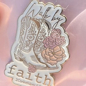 May include: A gold-rimmed enamel pin featuring white cowboy boots with floral details, pink flowers, and the text Walk by faith 2Corinthians 5:7. The design is set against a white background with gold metal outlines and lettering.