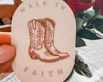 May include: A pink oval sticker with the text "Walk in Faith" in brown lettering. The sticker features a pair of brown cowboy boots with floral designs.