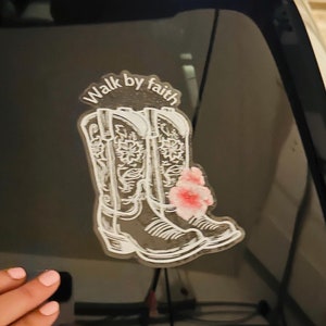 May include: A white vinyl sticker with the text "Walk by faith" and a pair of white cowboy boots with pink flowers on the back of a car window.