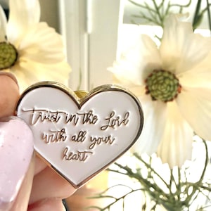 May include: A heart-shaped enamel pin with a white background and gold-colored metal trim. The pin features the engraved text "Trust in the Lord with all your heart" in a cursive script font, centered on the white surface of the heart.