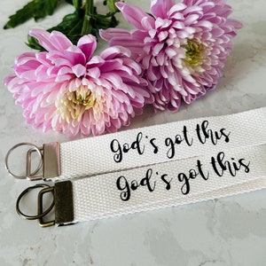 May include: Two white canvas keychains with black text that reads "God's got this". The keychains are lying on a white surface with pink flowers.