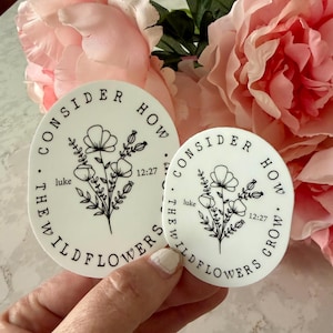 May include: Two white circular stickers with black text and floral illustrations. The text reads "CONSIDER HOW THE WILDFLOWERS GROW" and "luke 12:27". The stickers are held in front of pink flowers.