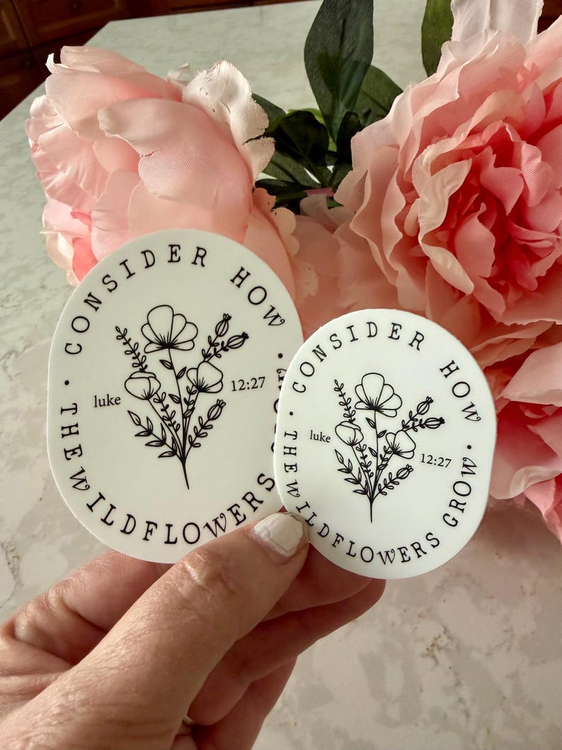 May include: Two white circular stickers with black text and floral illustrations. The text reads "CONSIDER HOW THE WILDFLOWERS GROW" and "luke 12:27". The stickers are held in front of pink flowers.