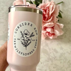 May include: A light pink tumbler with a silver rim and a matching pink straw. The tumbler has a white sticker with a floral design and the text "CONSIDER HOW WILDFLOWERS GROW" and "luke 12:27". Pink flowers are in the background.