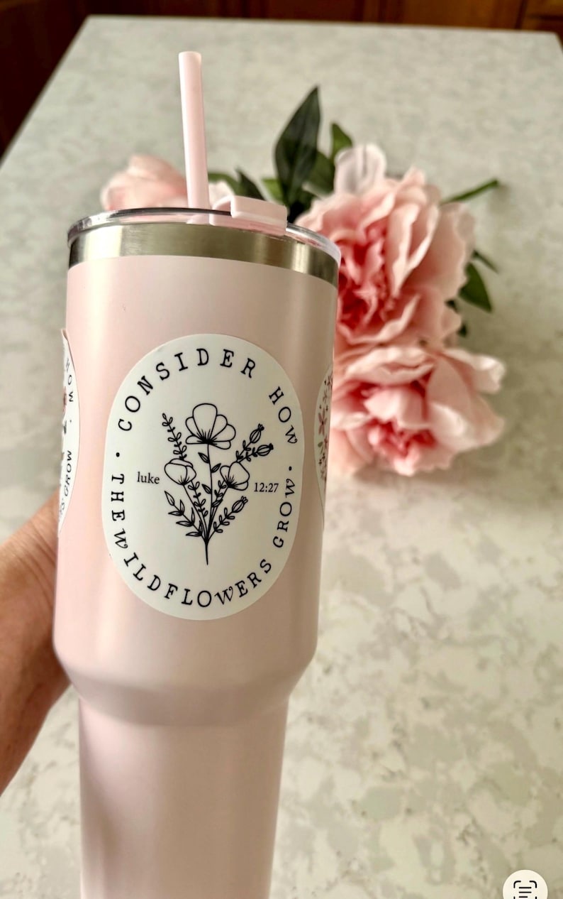 May include: A light pink tumbler with a silver rim and a matching pink straw. The tumbler has a white sticker with a floral design and the text "CONSIDER HOW WILDFLOWERS GROW" and "luke 12:27". Pink flowers are in the background.