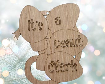 May include: A light brown wooden ornament shaped like a cluster of three Christmas light bulbs. The text "It's a beaut Clark" is engraved across the surface of the bulbs in a rounded, playful font. A small hole is at the top for hanging.