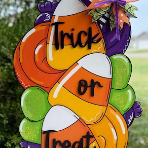 May include: A wooden Halloween decoration shaped like candy corn with the words "Trick or Treat". The decoration features orange, yellow, and white candy corn, green and purple accents, and a decorative bow.